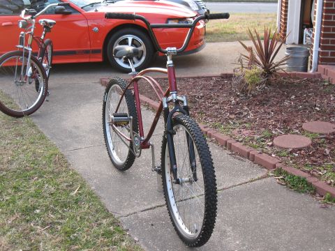 1994 Ford Bronco - A custom bicycle with a unique frame design and knobby tires is parked on a sidewalk, with another bicycle and a red car in the background.