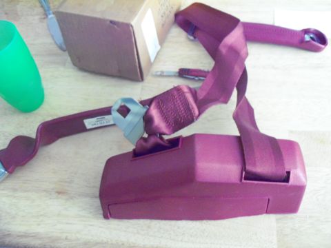 1979 Ford Ranchero - A maroon seatbelt assembly, including the buckle and housing, is shown on a wooden surface. Text is visible on the seatbelt strap.