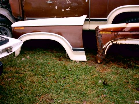 1980 Ford Bronco - Two brown-and-white front fenders removed and standing on the grass beside a truck body, the left one intact with amber marker light and ch…