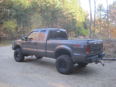 2003 Ford F-350 - A lifted 2003 Ford F-350 Super Duty pickup truck is shown from a 3/4 front angle on a dirt road with trees in the background.