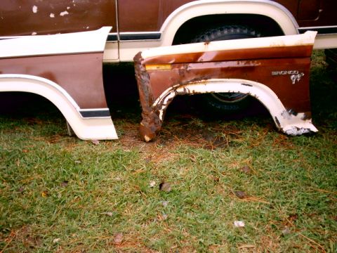 1980 Ford Bronco - Two removed brown-and-white front fenders leaned against a truck on grass, the right one badly rusted through behind the wheel opening and…