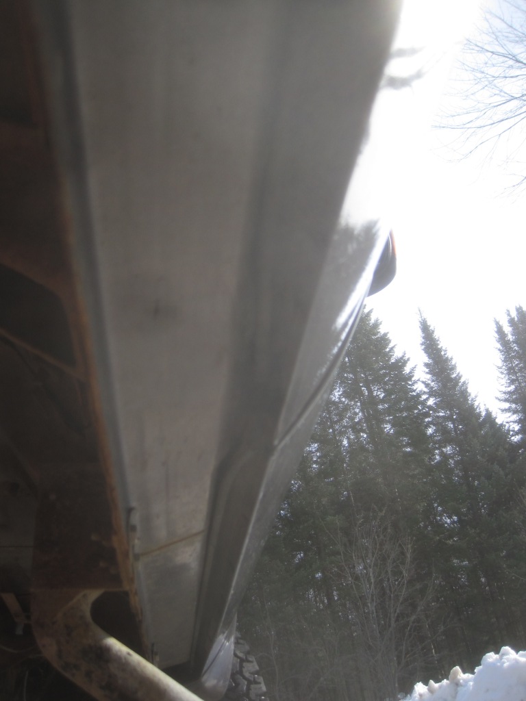 Truck body panel with visible rust and a curved edge, seen from a low angle against a backdrop of trees