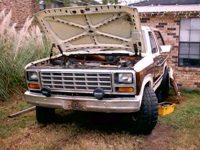 Brown and tan Ford Bronco with hood open in a yard, engine bay exposed above chrome grille