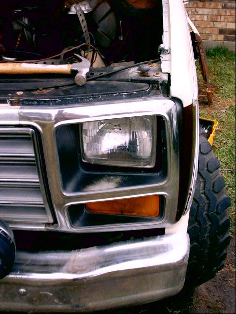 1980 Ford Bronco - Close-up of the driver-side front corner of a white Ford truck with the hood open, showing the rectangular sealed-beam headlight, amber tur…