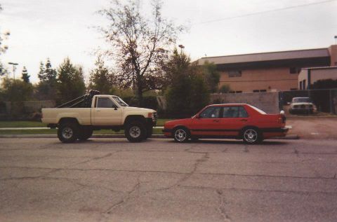 1988 Toyota Pickup/SR5 - A lifted Toyota pickup truck is parked next to a red sedan on a street with buildings in the background.