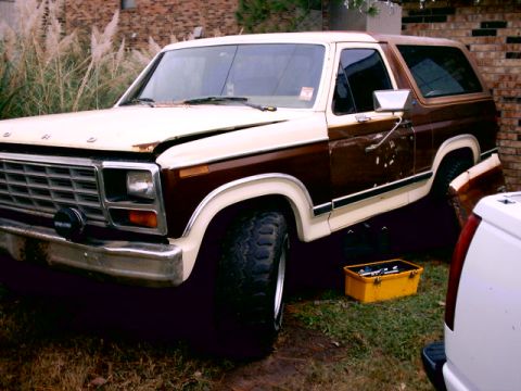 1980 Ford Bronco - Front 3/4 view of a brown and tan two-tone full-size Ford Bronco parked on grass beside a brick house, with a faded hood, dented driver's d…