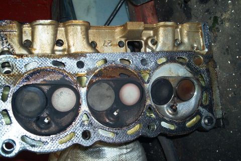 1993 Ford Taurus - Removed cylinder head laid face-up, showing three two-valve combustion chambers, two carbon-fouled and one steam-cleaned bright and clean,…