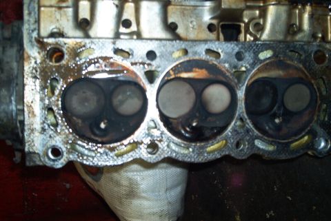 1993 Ford Taurus - Removed aluminum cylinder head resting face-up on a rag, showing three combustion chambers each with two valves, carbon-blackened chamber w…