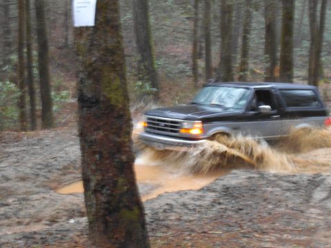 1986 Ford Bronco - A Ford Bronco navigates a muddy trail in a wooded area, kicking up water.