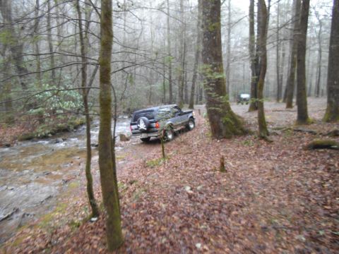 1986 Ford Bronco - A dark SUV navigates a wooded area with a stream beside the path.