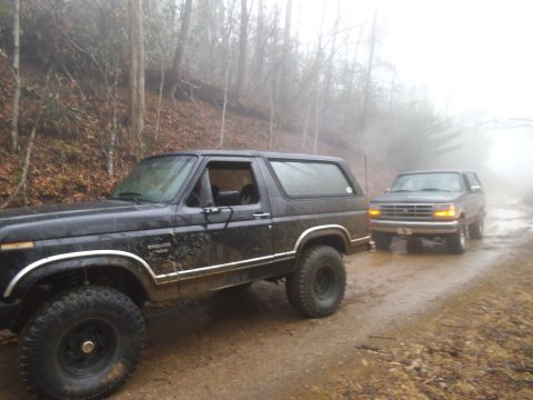 1986 Ford Bronco - Two Ford Broncos drive down a muddy, foggy dirt road surrounded by trees.