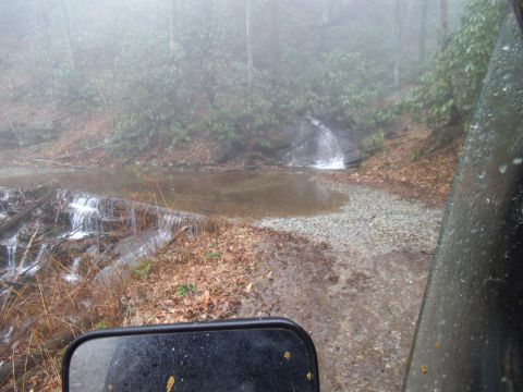1986 Ford Bronco - View from inside a vehicle as it approaches a stream crossing in a foggy, wooded area.