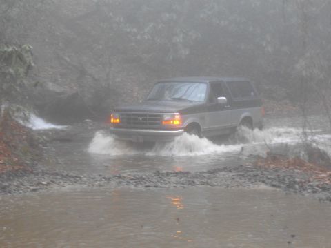 1986 Ford Bronco - A two-tone Ford Bronco navigates a flooded path, kicking up water.