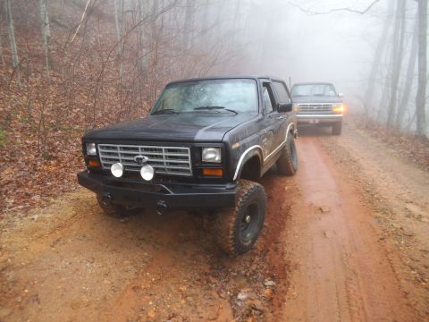 1986 Ford Bronco - Two SUVs drive down a muddy, wooded road on a foggy day.