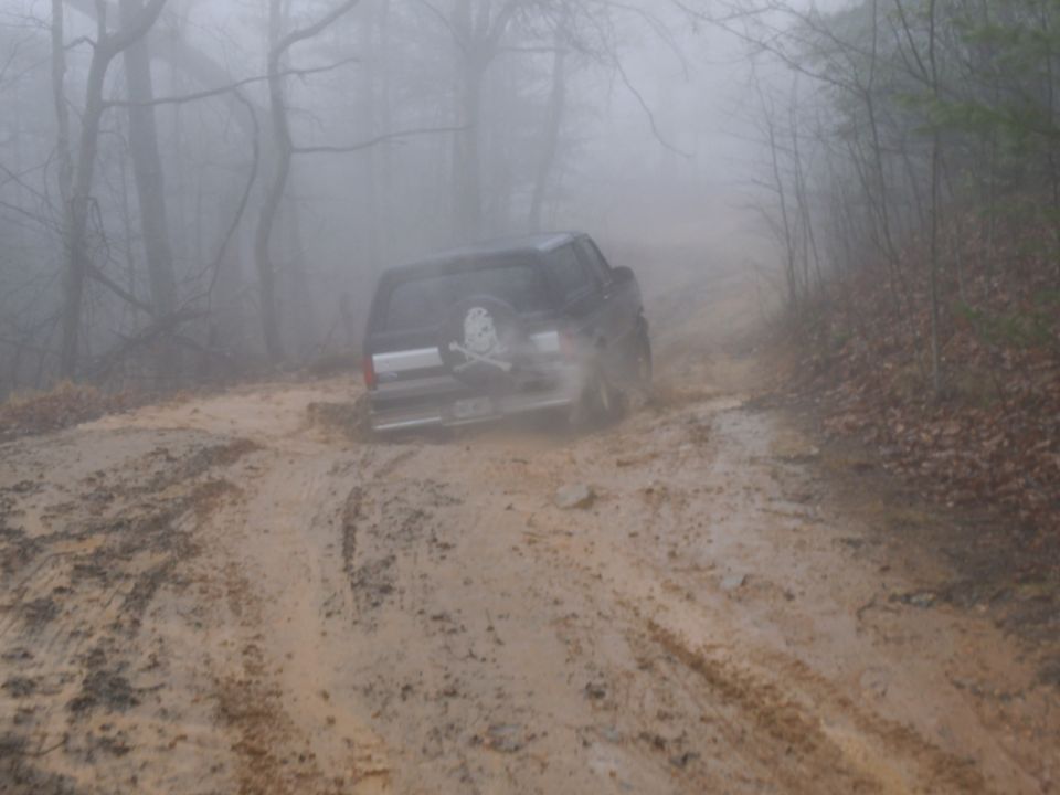 Black 1986 Ford Bronco driving through a muddy forest road
