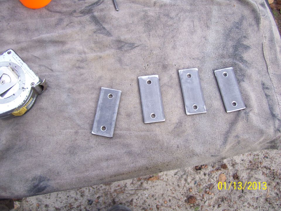 Four metal brackets with two holes each, laid out on a cloth next to a tape measure