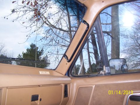1990 Ford Bronco - Interior corner of a tan-trimmed truck showing the A-pillar, opening vent window and the side mirror with bare trees outside.