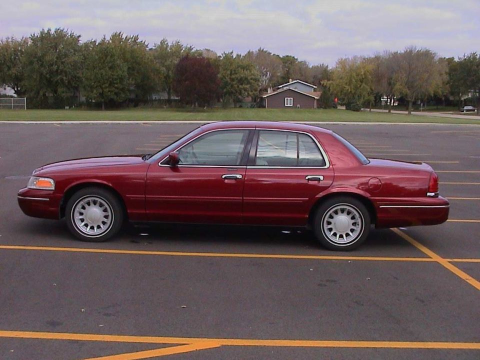 Dark red Ford Crown Victoria sedan, driver-side profile, parked in an empty lot with trees behind