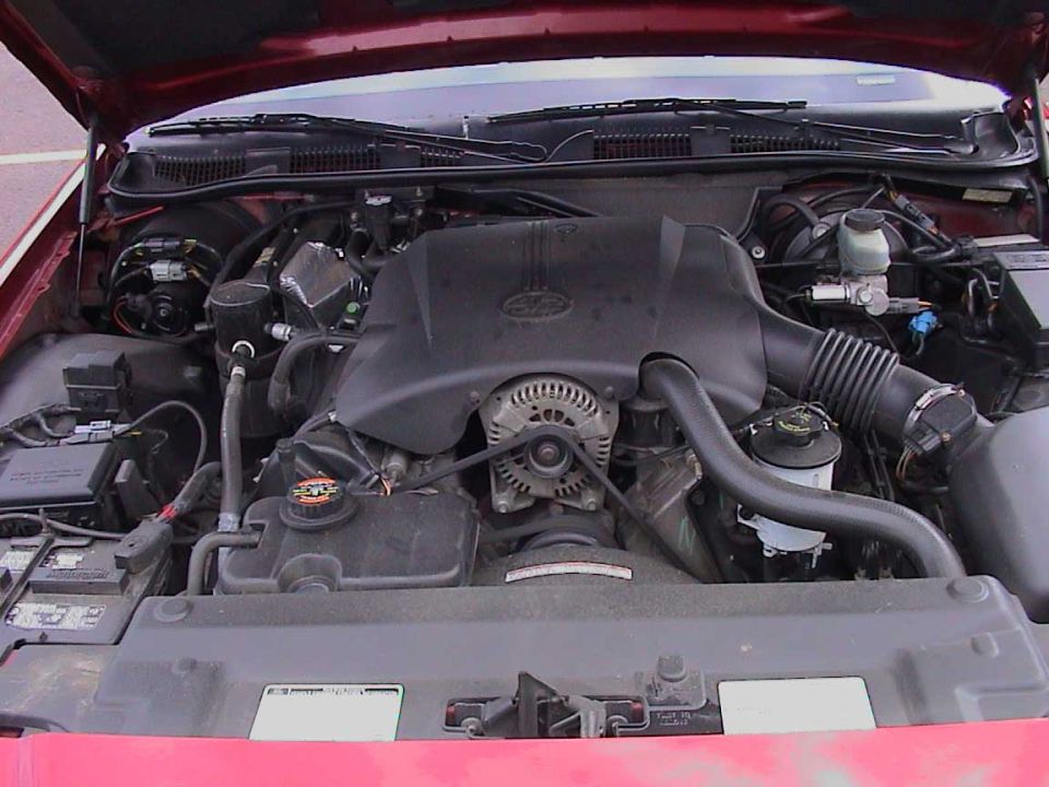 Engine bay of a dark red sedan showing V8 with black intake cover, alternator, and air intake tube