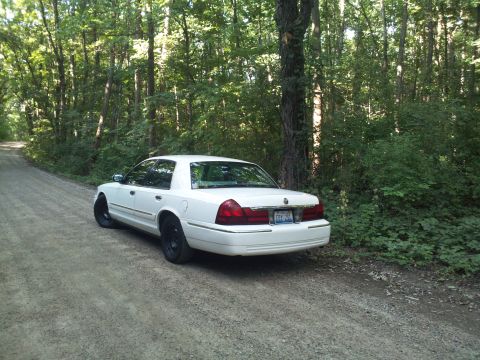 2000 Mercury Grand Marquis - A white sedan is parked on a dirt road surrounded by trees. The rear of the vehicle is facing the camera.