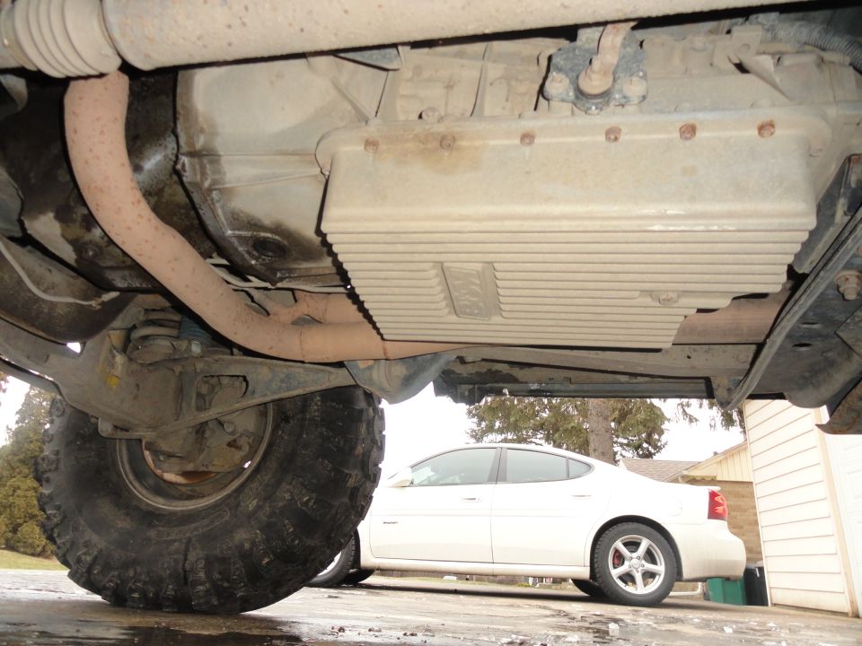 Undercarriage view showing large off-road tires and transmission pan, with a white sedan in the background