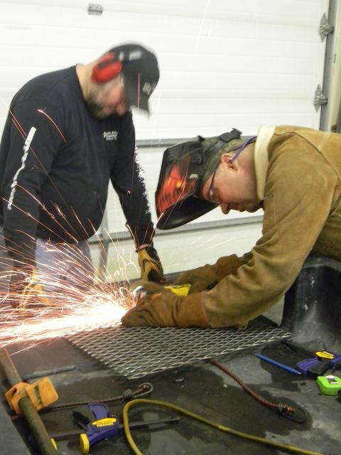 1990 Chevrolet Suburban - Two men are working on a metal grate with a grinder, creating a shower of sparks in a garage setting.