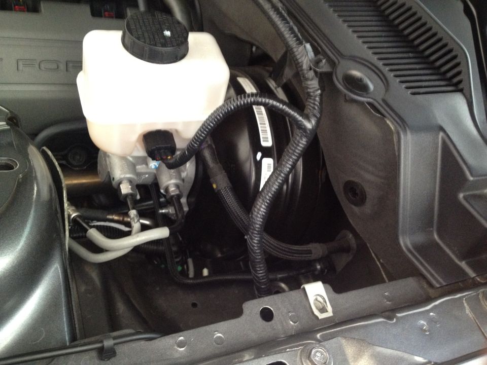 Close-up of the brake master cylinder and booster assembly in a 2013 Ford Mustang engine bay. Some wiring and hoses are visible