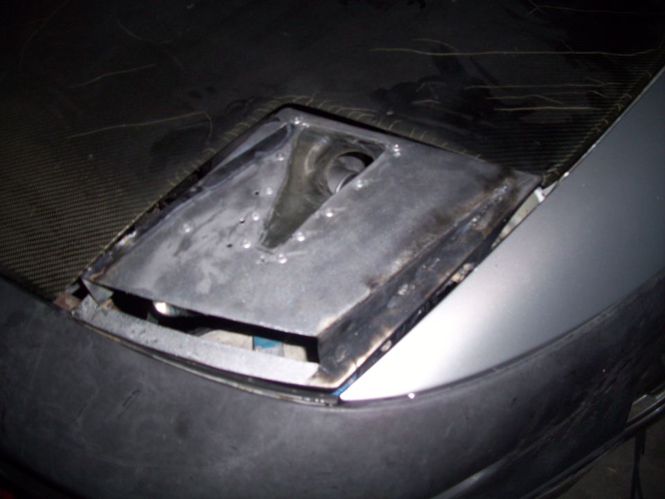close-up of a car's headlight housing, appears to be modified or under repair, with a carbon fiber hood visible