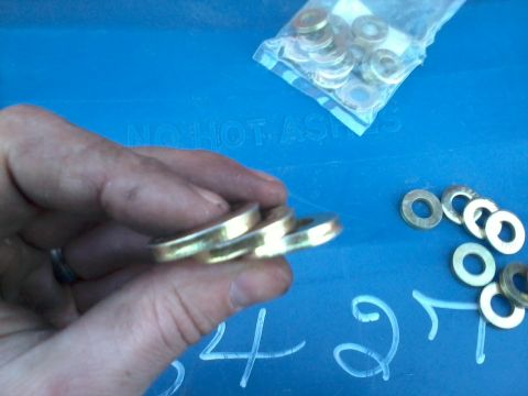 1981 Ford Bronco - A hand holds three brass washers, with a bag of washers and loose washers scattered on a blue surface with handwritten numbers.