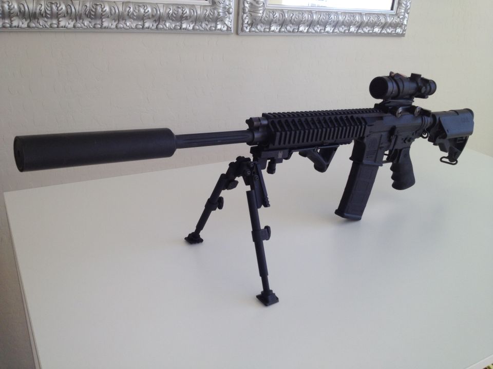 Black rifle with bipod, scope, and silencer on a white surface