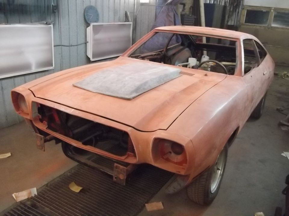 Car body in primer, looks like a 1974 Ford Mustang II, hood scoop, in a workshop