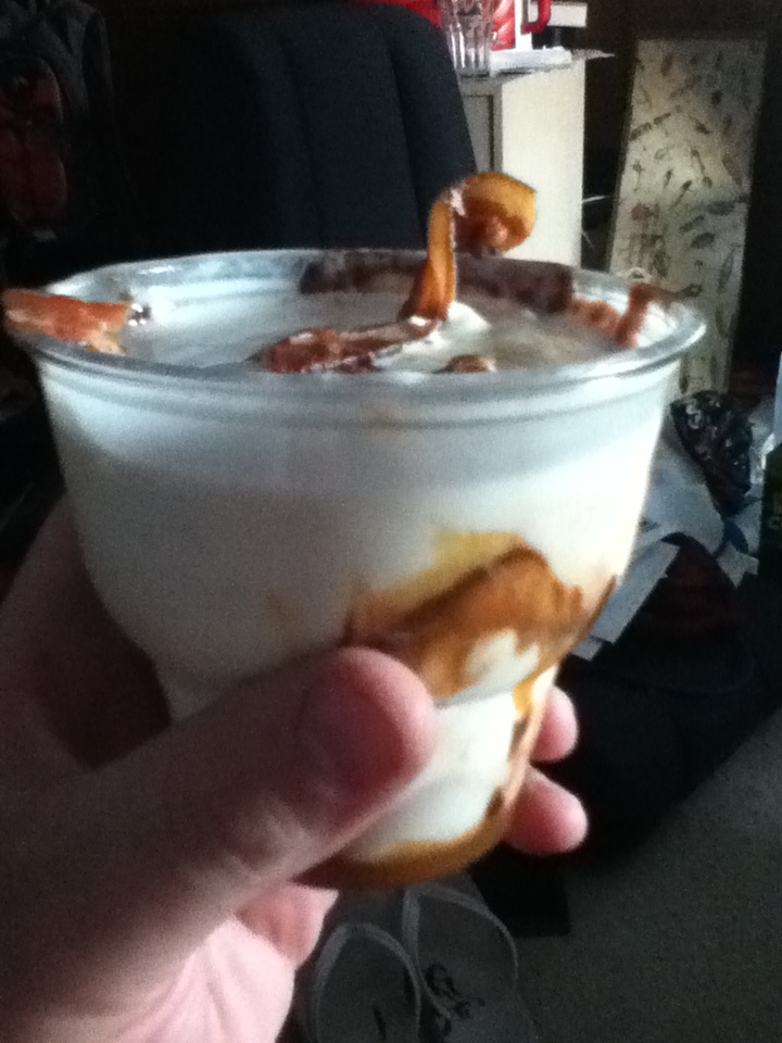 Hand holding a clear cup with ice cream and caramel sauce, topped with what looks like a cookie or pastry piece sticking out