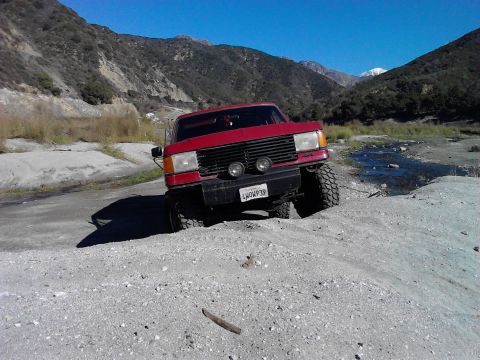 1988 Ford Bronco - This red SUV is navigating a rough, sandy terrain with mountains in the background.
