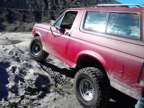 1988 Ford Bronco - This red SUV is navigating a rough, sandy terrain with large off-road tires.