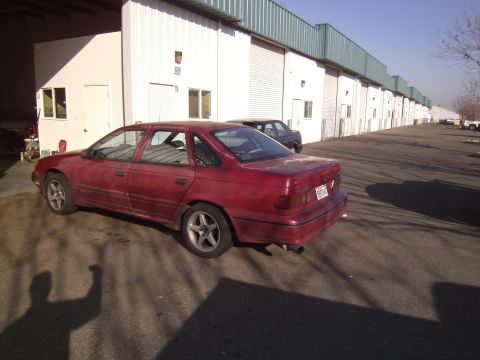 1991 Ford Taurus - A red 1991 Ford Taurus sedan is parked in front of a row of industrial buildings with a second dark car behind it.