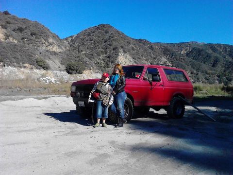 1988 Ford Bronco - A red SUV is parked on a dirt lot with two people standing in front of it, with mountains in the background.
