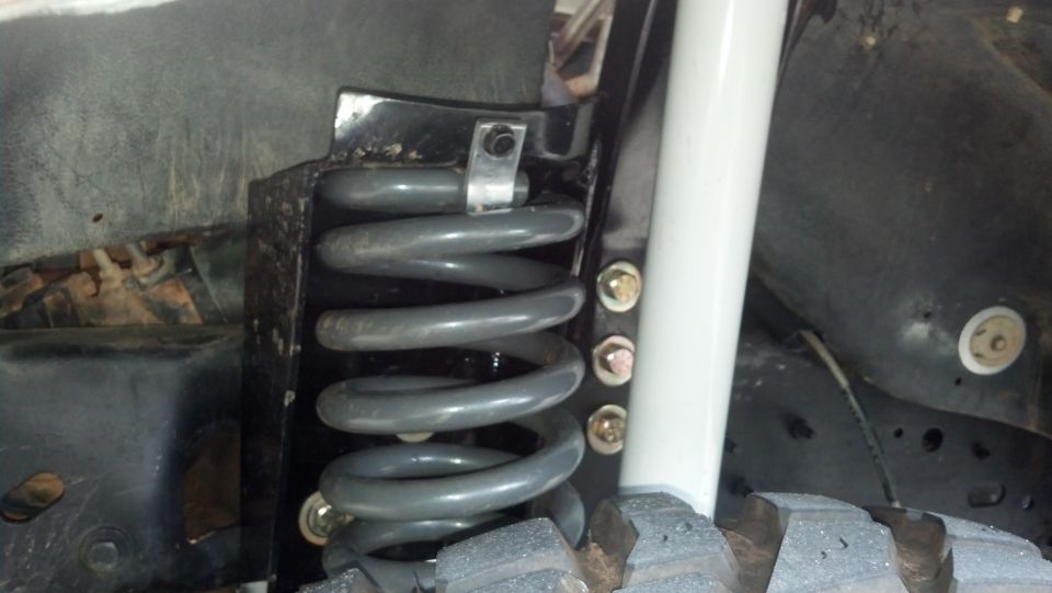 Gray coil spring in bucket with bolted drop bracket above mud-terrain tire
