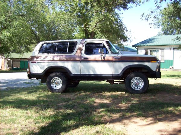 White and brown 1978 Ford Bronco side view, lifted with big tires, parked on grass near a house