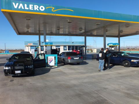 2003 Ford Crown Victoria - Several dark sedans fueling under a Valero station canopy as two men stand beside a car, desert beyond