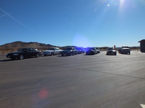 2003 Ford Crown Victoria - Row of dark and silver sedans plus a pickup parked across a paved desert lot with hills in the distance