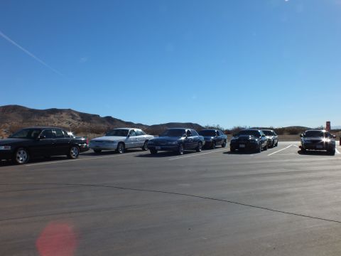 2003 Ford Crown Victoria - Line of black and silver four-door cars angled across a desert lot, dry hills in the background