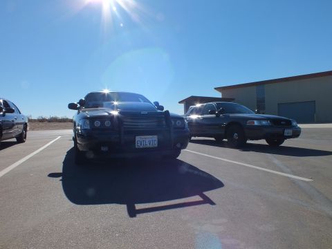 2003 Ford Crown Victoria - EVIL VIC Charger parked alongside a Ford Crown Victoria near a metal warehouse