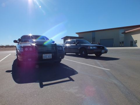 2003 Ford Crown Victoria - Charger with EVIL VIC plate and a sedan sit on pavement near a tan warehouse