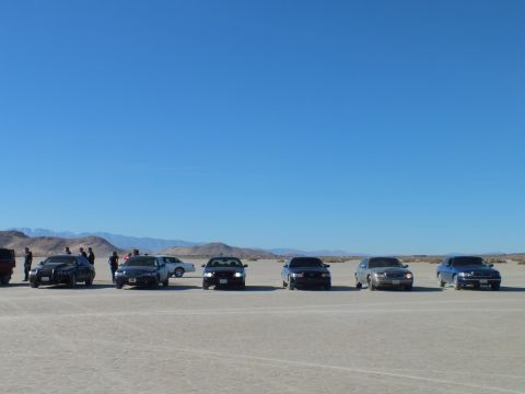 2003 Ford Crown Victoria - Six sedans with lights on spaced across a flat dry lake under clear sky