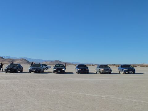 2003 Ford Crown Victoria - Row of sedans across a desert dry lake as people gather near the left vehicles