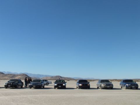 2003 Ford Crown Victoria - Lineup of dark and silver sedans on a flat dry lake with people at the left