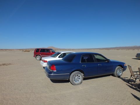2003 Ford Crown Victoria - Blue sedan and a red SUV on a desert dry lake with a folding chair at right
