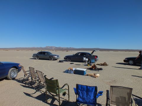 2003 Ford Crown Victoria - Sedans gathered on a flat desert lakebed; empty camp chairs and a cooler sit in the sandy foreground under blue sky.