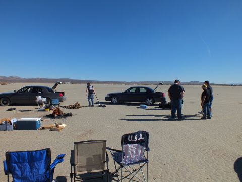 2003 Ford Crown Victoria - Group works near two dark sedans with raised trunks on a desert flat; a USA-flag camp chair sits in the foreground.