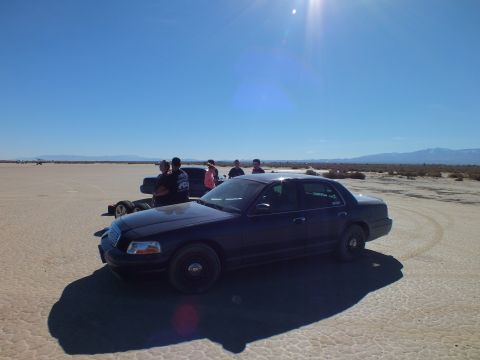 2003 Ford Crown Victoria - Dark Crown Victoria sedan sits on a desert flat as people stand behind it; strong sun flare crosses the frame.